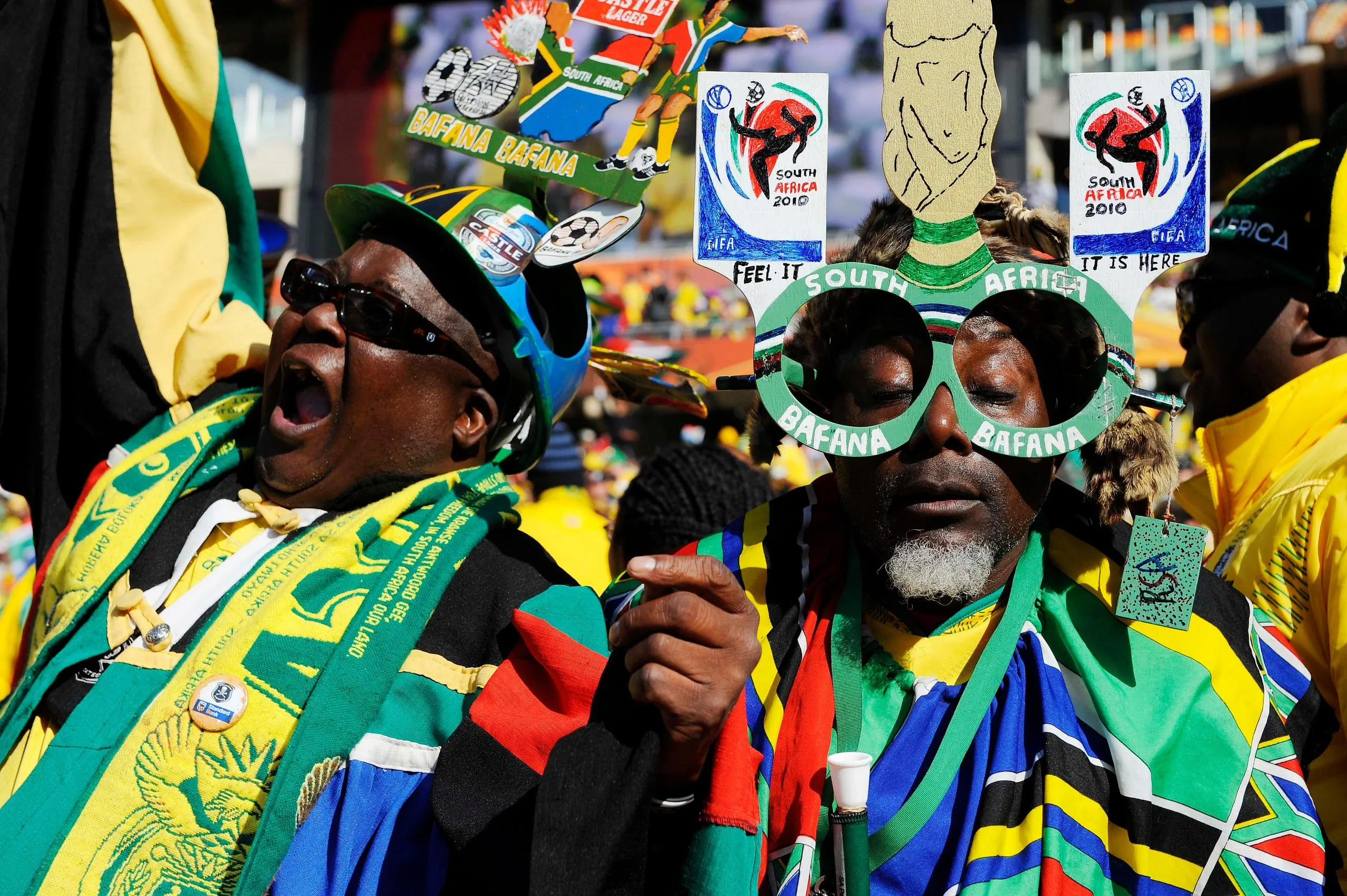 Two South Africa fans in team shirts and scarves, one wearing a home-made hat and one wearing home-made glasses, cheer for their team at the 2010 World Cup.